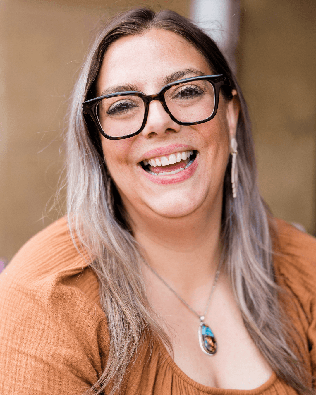 Mulher sorridente com óculos de armação preta, cabelos longos e blusa cor de terracota.