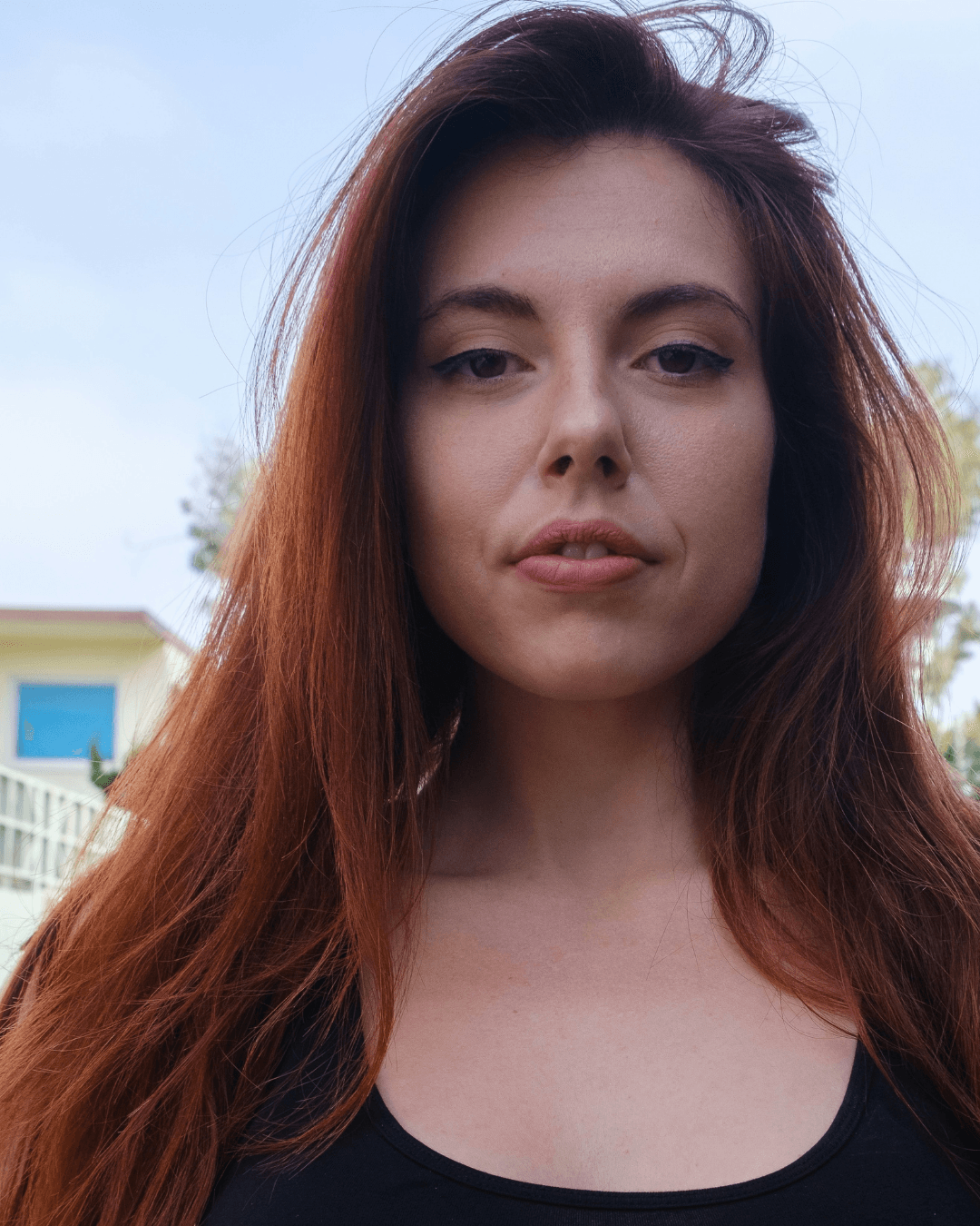 Mulher com longos cabelos ruivos, olhos escuros e batom rosado olhando para a câmera.